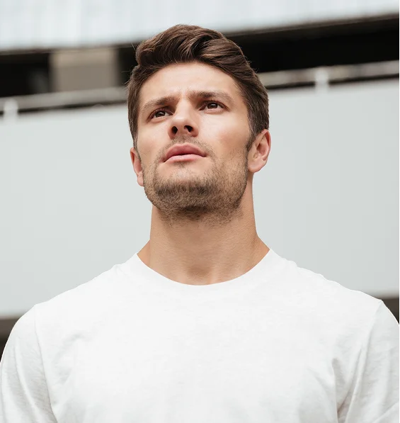 A close-up outdoor portrait of a handsome man in a white t-shirt, looking upward with a serious expression.