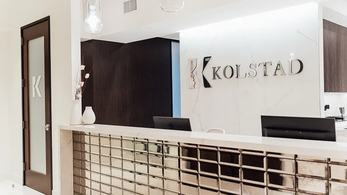 Close-up view of the KOLSTAD clinic's modern reception area, showing the metallic tiled counter, the brand logo on the marble wall, and a frosted glass door.