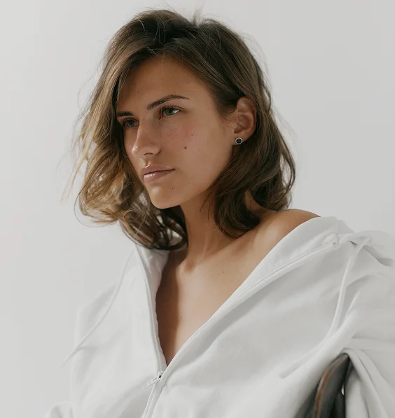 A young woman with brown, shoulder-length hair looks to the left, with her profile visible. She is wearing a white hoodie or top that is pulled down to expose her shoulder, and her hand is resting near her neck, gently touching her hair. She has a dark stud earring in her ear. The background is a plain, light color.