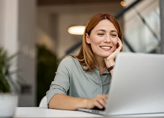 A smiling woman with red-brown hair sitting at a desk, looking happily at the camera while working on a silver laptop.