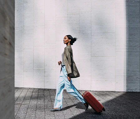 A stylish woman in a business jacket and light blue trousers walking across a sunlit, paved area while pulling a red rolling suitcase.