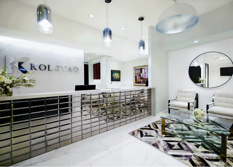 The bright, modern, and luxurious reception area of the KOLSTAD clinic, featuring a shiny chrome-tiled desk, marble floor, and comfortable white seating.