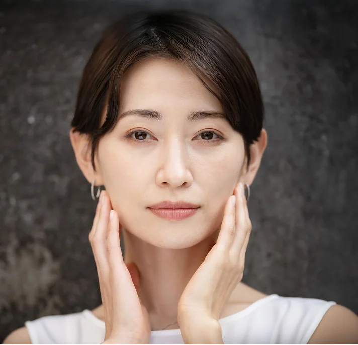 A portrait of an East Asian woman with dark hair pulled back, touching both sides of her face, looking directly at the camera with a neutral expression.