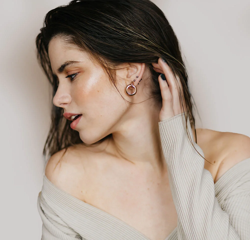 Profile portrait of a woman with dark, wet-look hair, wearing an off-the-shoulder grey sweater and touching her ear.