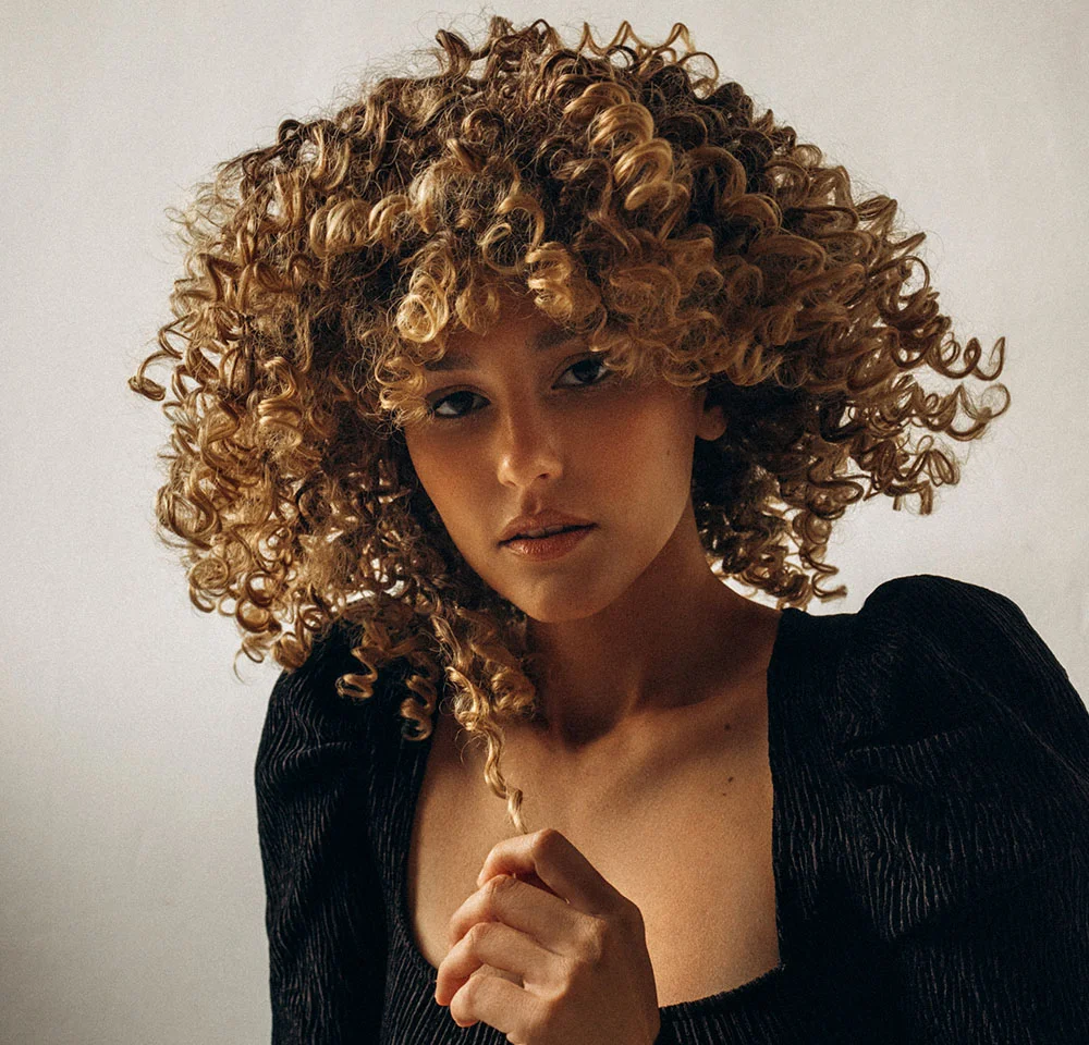 Headshot of a woman with voluminous, light brown curly hair, wearing a black top with puffy sleeves, looking intensely at the camera.
