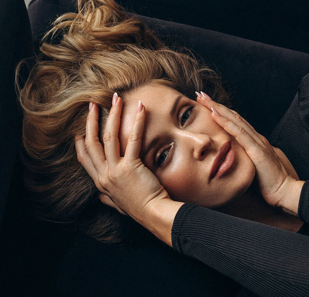 Close-up portrait of a woman with light blonde hair, holding her hands on her face while lying down against a dark background.