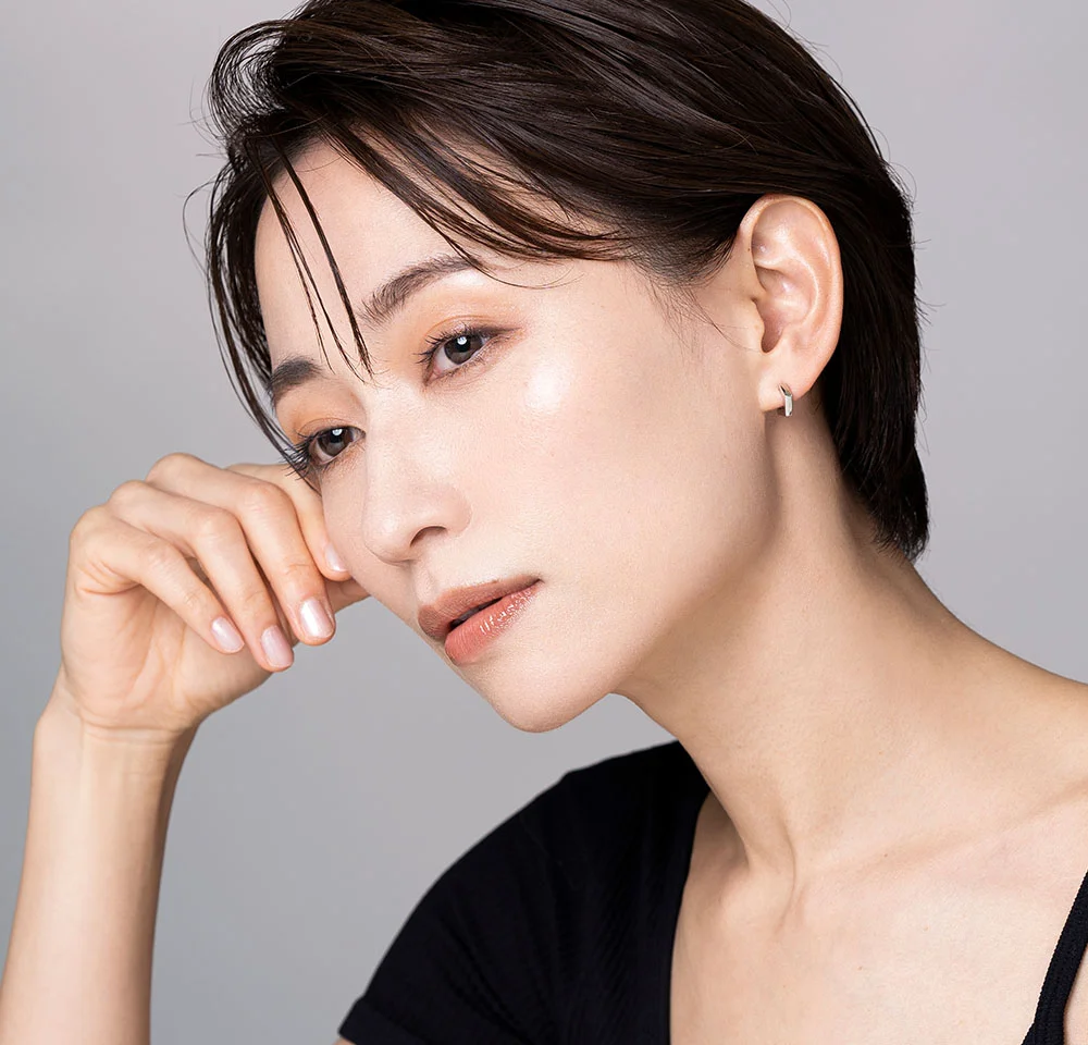 Close-up portrait of a woman with short, wet-look dark hair, wearing a black top and gently touching her cheek.
