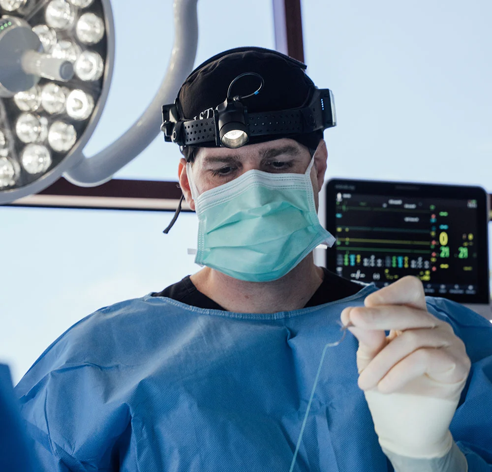 Close-up of Dr. Kolstad in surgical attire and mask, holding a surgical thread or suture material with gloved hands, focused on the task.