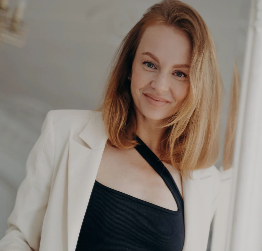 Close-up portrait of a smiling woman with medium reddish hair, wearing a white blazer over a black top with a cutout.