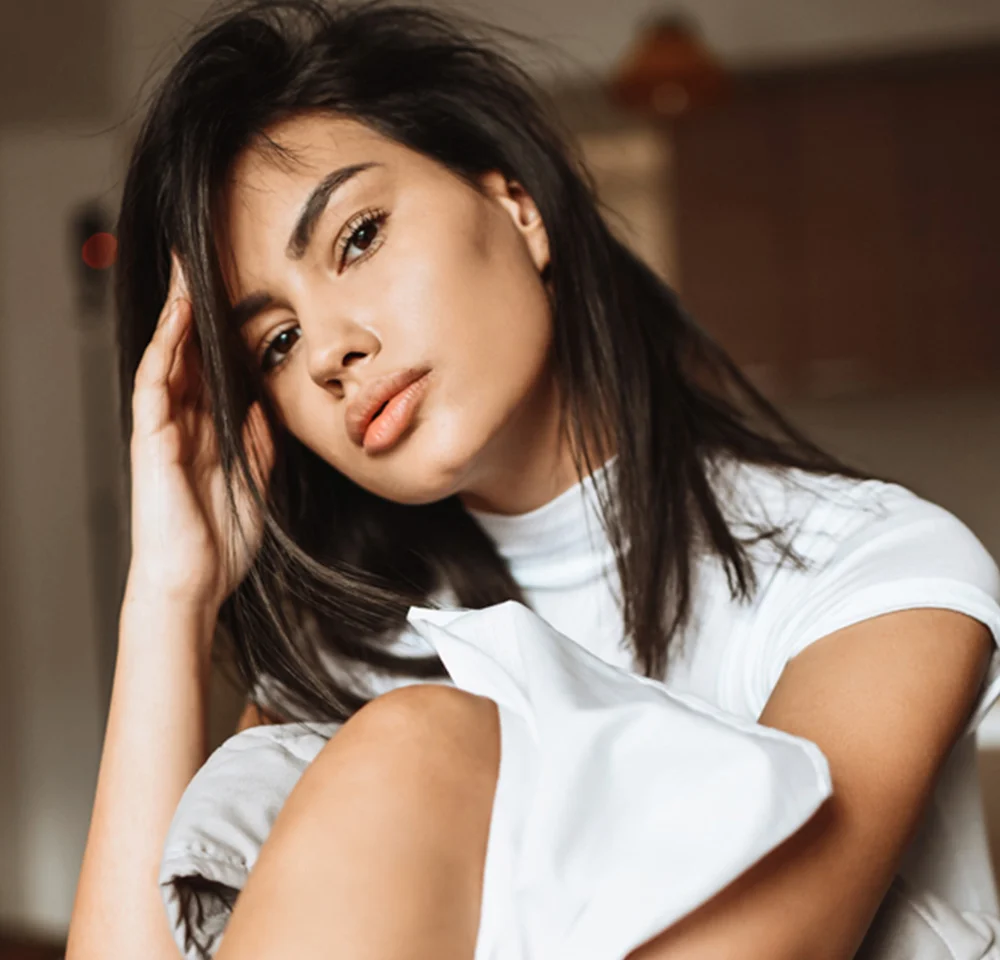 A beautiful young woman with dark hair and a white top posing indoors, with dramatic lighting. Focuses on her cheek and jawline definition.