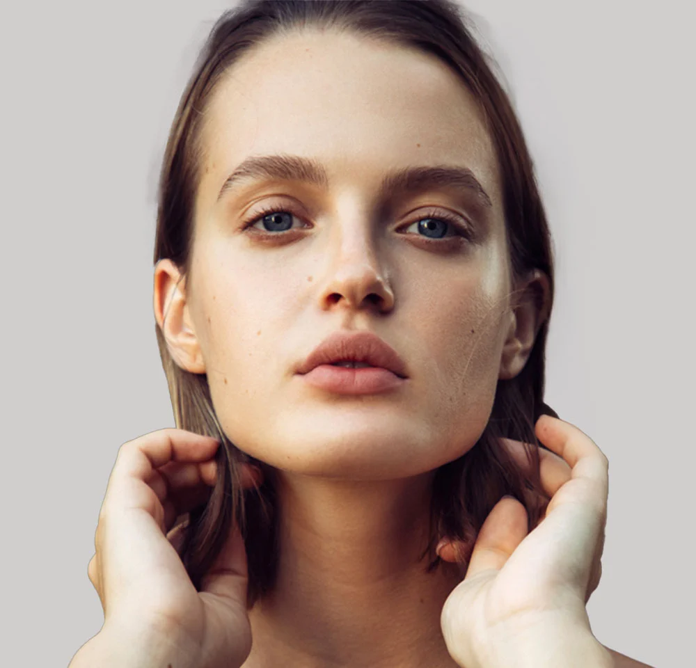 Headshot of a woman with light brown hair and blue eyes, looking directly at the camera while holding her hands near her jawline.