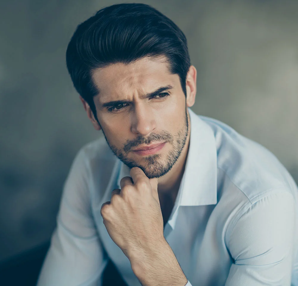 Headshot of a man in a light blue collared shirt, with a serious expression and his hand resting on his chin, looking away from the camera.