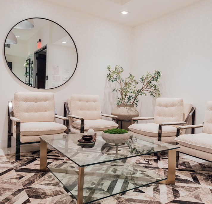 Interior photo of a stylish waiting area featuring four cream-colored chairs with metal frames, a glass coffee table, and a large circular mirror on the wall.