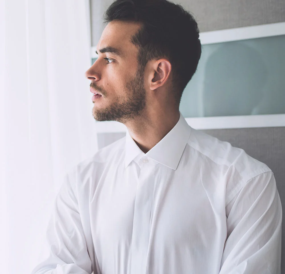 Profile shot of a man in a white collared shirt, with dark hair and a short beard, looking out a window.