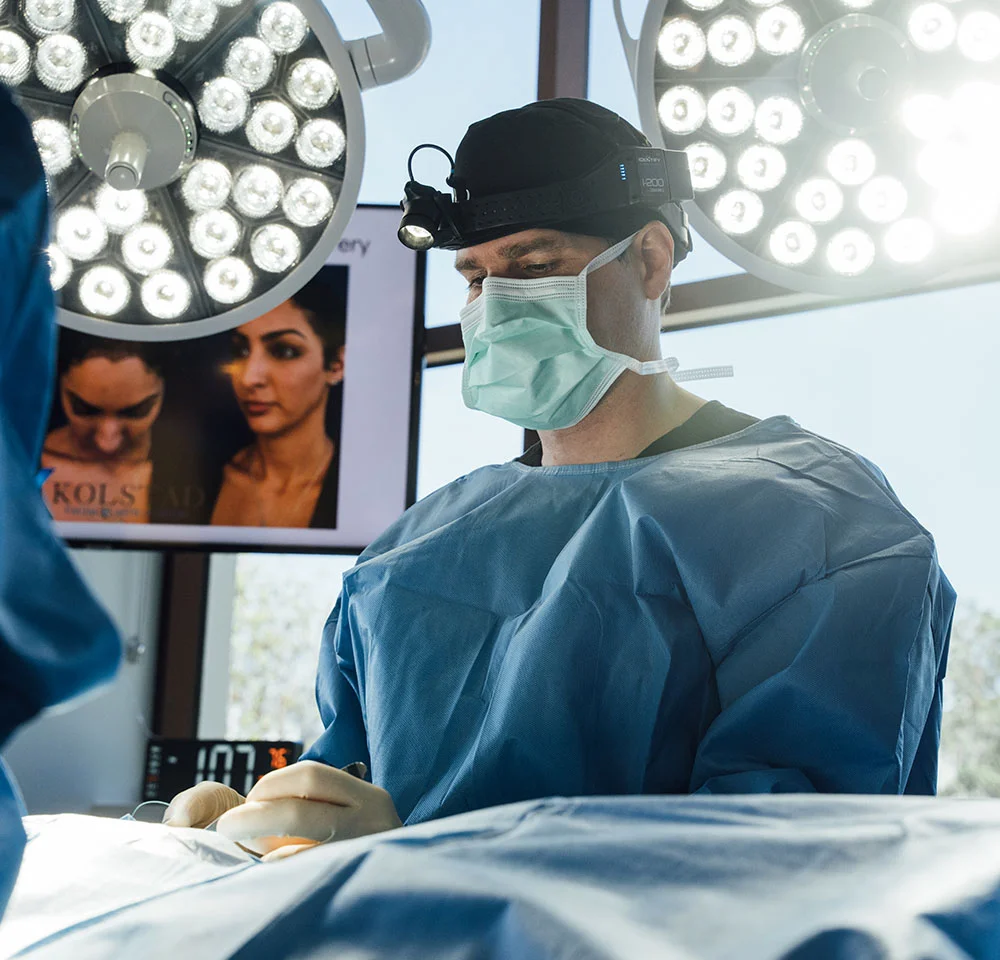 Dr. Kolstad performing surgery; a monitor behind him shows before-and-after photos labeled 'KOLSTAD' related to Middle Eastern Rhinoplasty.