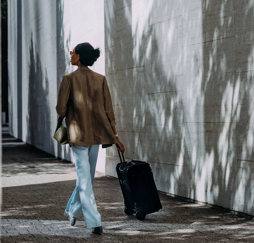 A woman wearing a brown blazer and light blue pants walks away from the camera, pulling a black roller suitcase along a stone walkway with striped shadow patterns.