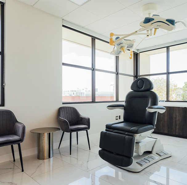 Interior of a modern medical or treatment room featuring a large, black leather electric procedure chair in the center, two gray velvet side chairs, a small metallic side table, and large windows overlooking a city view. Operating lights hang from the ceiling.