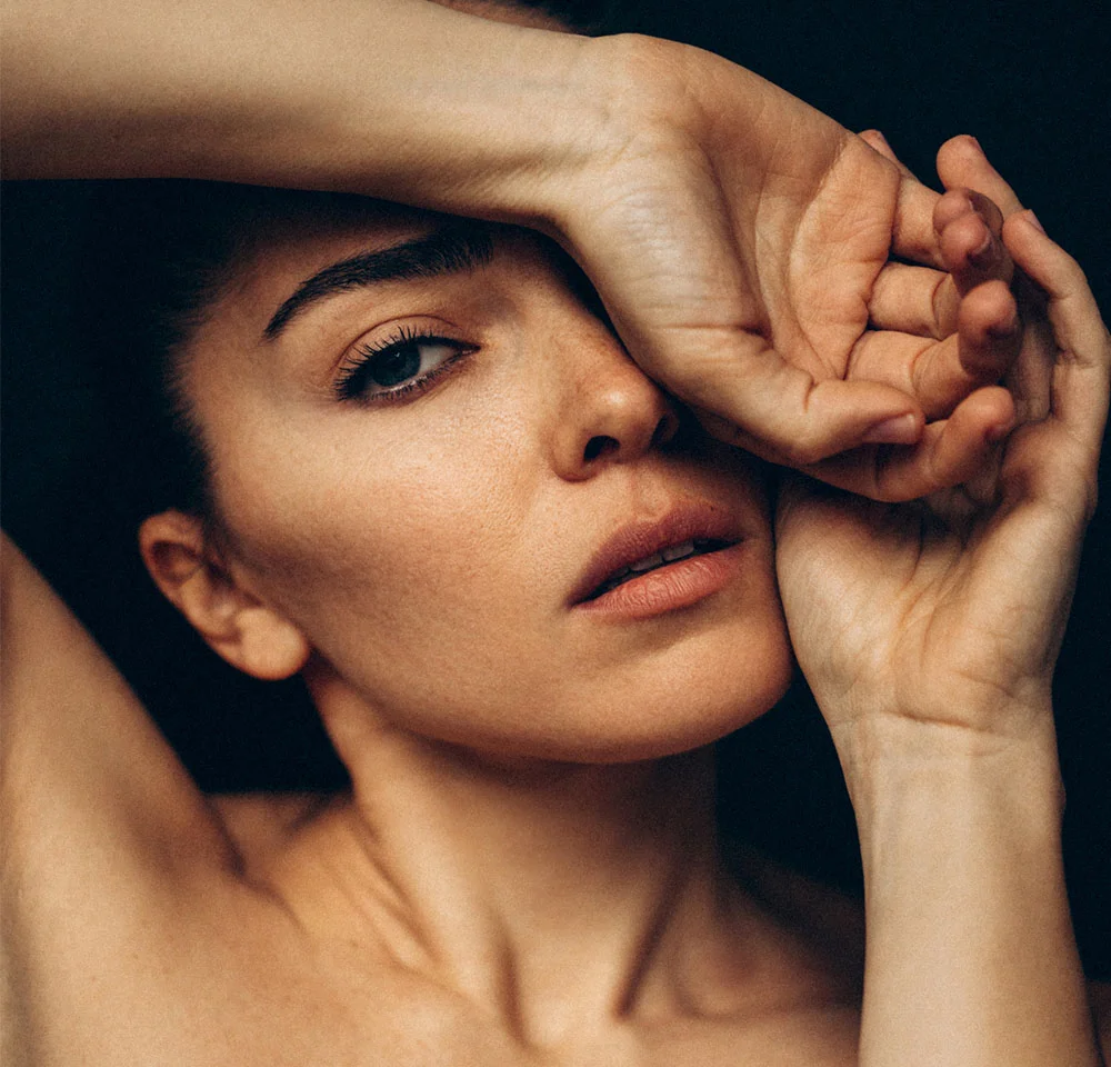 A close-up, dramatic portrait of a person with dark hair, looking intently at the camera while covering their forehead with one hand and touching their cheek with the other against a dark background. - Under Eye Fillers in La Jolla, CA